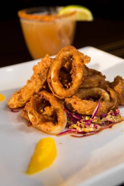 Güzel altın gevrek derin kızarmış kalamar halkaları, turuncu kesimleri ve arka planda yak²n bir kokteyl ile beyaz bir tabakta servis.