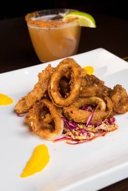 Güzel altın gevrek derin kızarmış kalamar halkaları, turuncu kesimleri ve arka planda yak²n bir kokteyl ile beyaz bir tabakta servis.