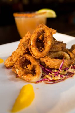 Güzel altın gevrek derin kızarmış kalamar halkaları, turuncu kesimleri ve arka planda yak²n bir kokteyl ile beyaz bir tabakta servis.