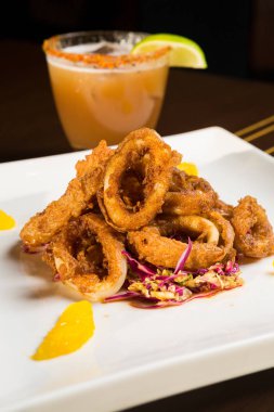 Güzel altın gevrek derin kızarmış kalamar halkaları, turuncu kesimleri ve arka planda yak²n bir kokteyl ile beyaz bir tabakta servis.