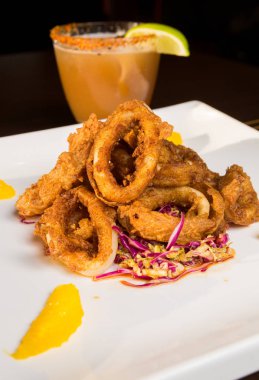 Güzel altın gevrek derin kızarmış kalamar halkaları, turuncu kesimleri ve arka planda yak²n bir kokteyl ile beyaz bir tabakta servis.