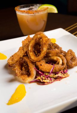 Güzel altın gevrek derin kızarmış kalamar halkaları, turuncu kesimleri ve arka planda yak²n bir kokteyl ile beyaz bir tabakta servis.