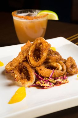 Güzel altın gevrek derin kızarmış kalamar halkaları, turuncu kesimleri ve arka planda yak²n bir kokteyl ile beyaz bir tabakta servis.