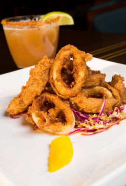 Güzel altın gevrek derin kızarmış kalamar halkaları, turuncu kesimleri ve arka planda yak²n bir kokteyl ile beyaz bir tabakta servis.