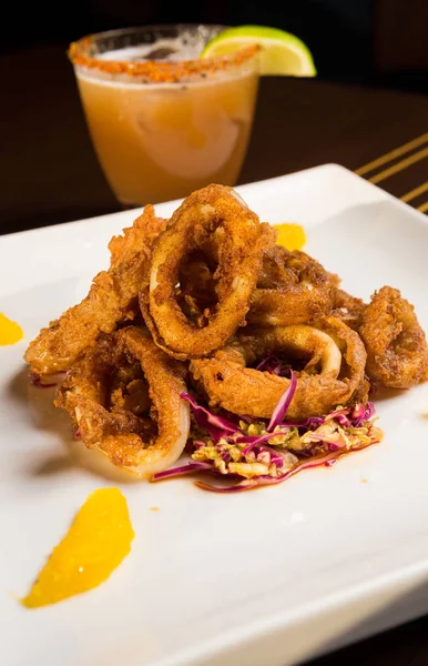 Güzel altın gevrek derin kızarmış kalamar halkaları, turuncu kesimleri ve arka planda yak²n bir kokteyl ile beyaz bir tabakta servis.