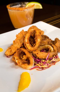 Güzel altın gevrek derin kızarmış kalamar halkaları, turuncu kesimleri ve arka planda yak²n bir kokteyl ile beyaz bir tabakta servis.