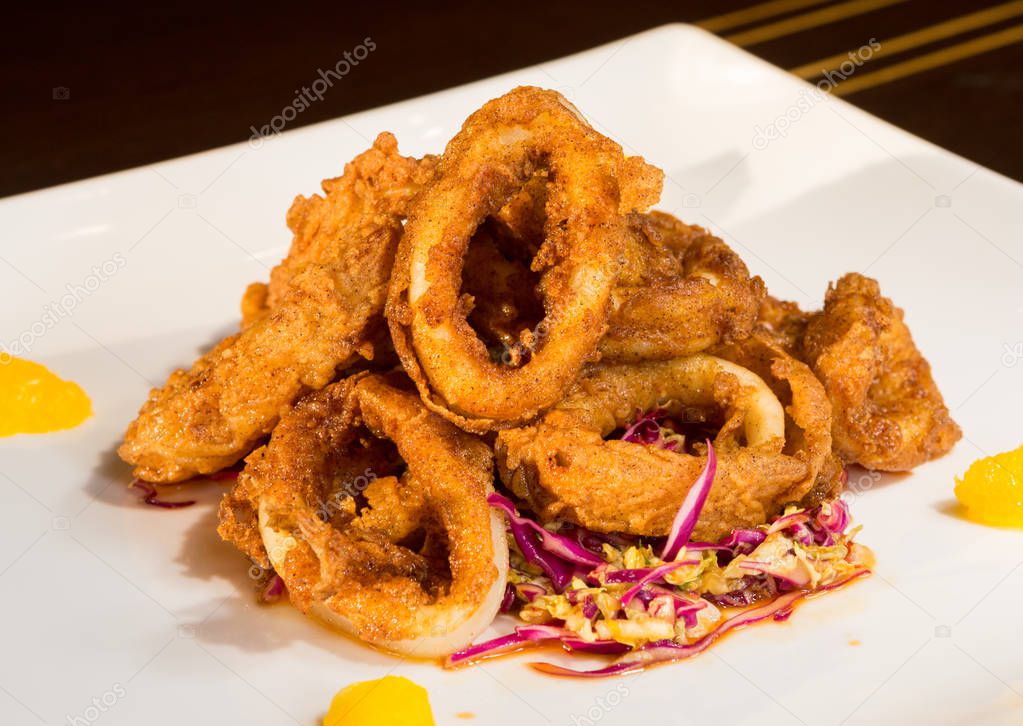 Beautiful golden crispy deep fried calamari rings, served on a white plate, with orange segments and a cocktail out of focus in the background.