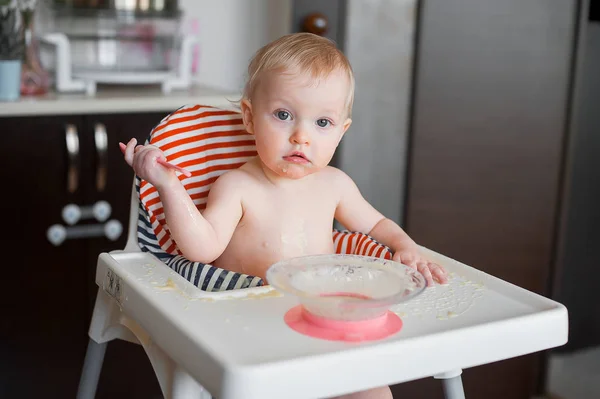 小可爱的女孩坐在高高的椅子和学习用勺子自己吃。有趣的快乐婴儿涂抹在桌子上的食物 — 图库照片