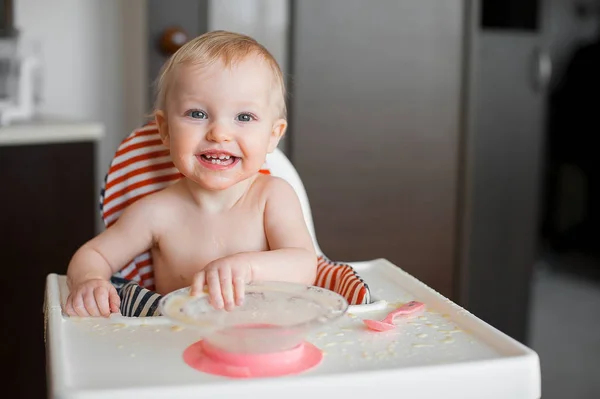 Μικρό χαριτωμένο κορίτσι κάθεται σε καρεκλάκι και μαθαίνει να τρώει με κουτάλι τον εαυτό της. Αστείο γέλιο ευτυχισμένο μωρό μουτζουρώματος φαγητό στο τραπέζι — Φωτογραφία Αρχείου