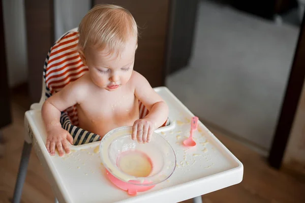 Μικρό χαριτωμένο κορίτσι κάθεται σε καρεκλάκι και μαθαίνει να τρώει με κουτάλι τον εαυτό της. Αστεία ευτυχισμένο μωρό μουτζουρώματος φαγητό στο τραπέζι — Φωτογραφία Αρχείου