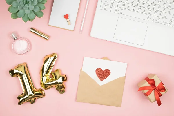 Flat lay home office desk. Women workspace with laptop, cosmetics, perfume, accessories on pink background. Love letter envelope, valentine red heart