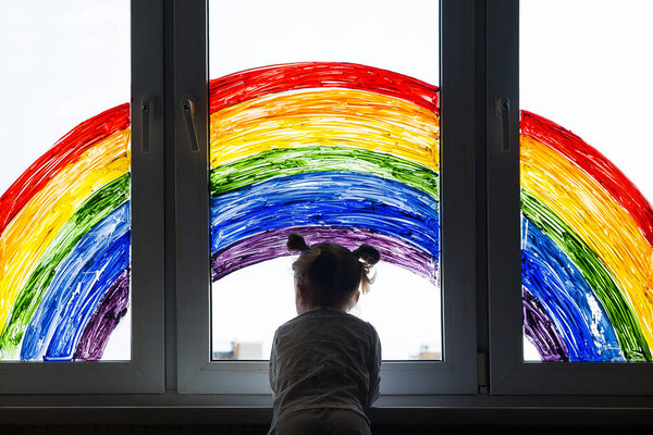 Little girl on background of painting rainbow on window. Kids leisure at home. Positive visual support during quarantine Pandemic Coronavirus Covid-19 at home. 