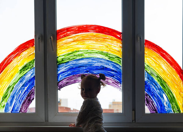 Little girl on background of painting rainbow on window. Kids leisure at home. Positive visual support during quarantine Pandemic Coronavirus Covid-19 at home. 