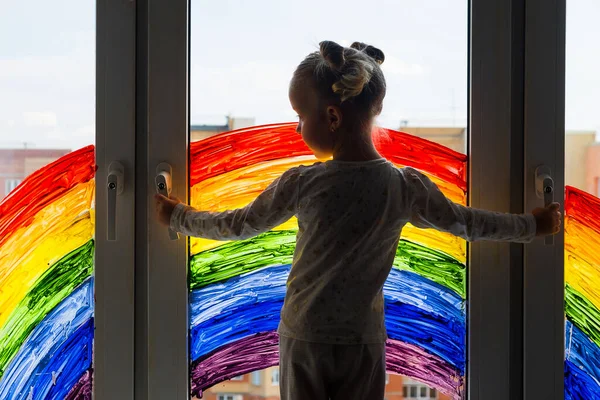 Little girl on background of painting rainbow on window. Kids leisure at home. Positive visual support during quarantine Pandemic Coronavirus Covid-19 at home.