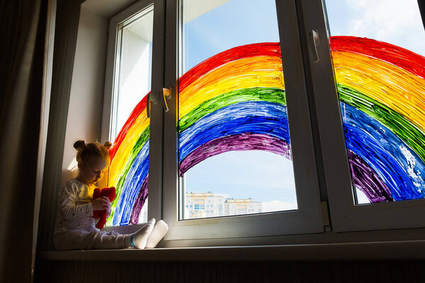 Little girl on background of painting rainbow on window. Kids leisure at home. Positive visual support during quarantine Pandemic Coronavirus Covid-19 at home. 