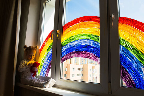 Little girl on background of painting rainbow on window. Kids leisure at home. Positive visual support during quarantine Pandemic Coronavirus Covid-19 at home. 