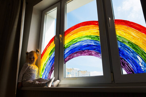Little girl on background of painting rainbow on window. Kids leisure at home. Positive visual support during quarantine Pandemic Coronavirus Covid-19 at home. 