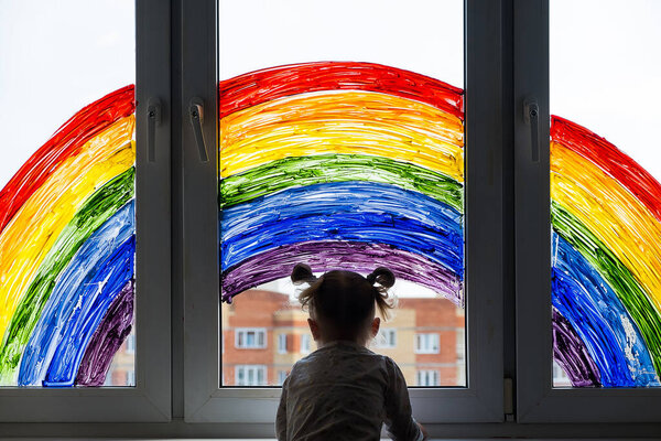 Little girl on background of painting rainbow on window. Kids leisure at home. Positive visual support during quarantine Pandemic Coronavirus Covid-19 at home. 