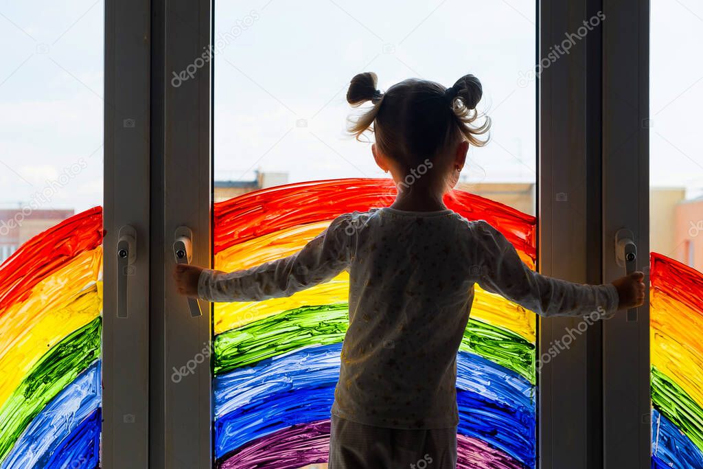 Little girl on background of painting rainbow on window. Kids leisure at home. Positive visual support during quarantine Pandemic Coronavirus Covid-19 at home. 