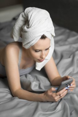 Kafasında havlu olan genç ve güzel bir kadın yatağında cep telefonunu tutuyor ve sosyal ağları kontrol ediyor. Uzaktan çalışma, online alışveriş, karantina tecrit