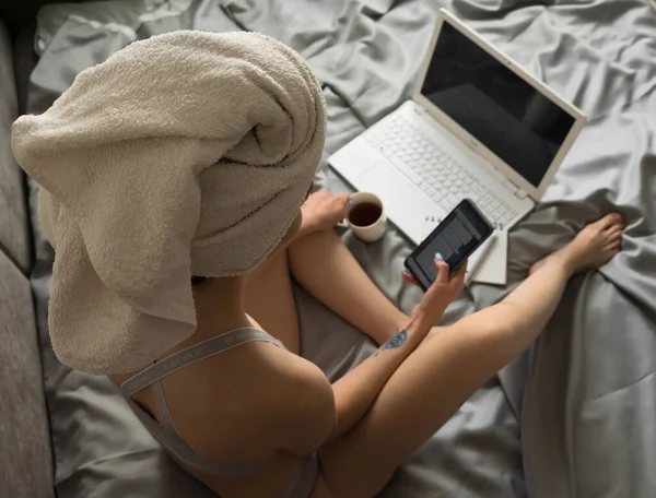 Csinos fiatal nő törülközővel a fején ül az ágyon, mobilt tart, és megnézi a közösségi oldalakat. Távmunka otthon laptoppal, online vásárlás, karantén elszigetelés. Felülnézet — Stock Fotó