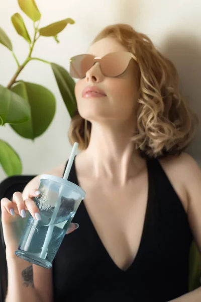 Young pretty woman holding fresh summer cocktail and looking away with smile while sitting at workspace, dreaming about vacation. Coronavirus situation in tourism industry. Quarantine. Isolation. — Stock Photo, Image