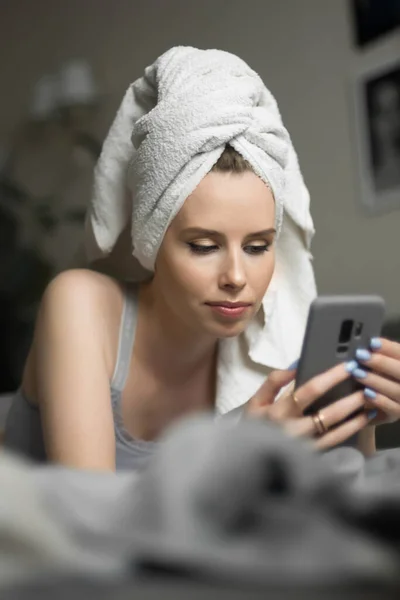 Bella giovane donna con un asciugamano in testa si trova sul telefono cellulare tenere a letto e controllare i social network. Lavoro a distanza, shopping online, isolamento in quarantena — Foto Stock