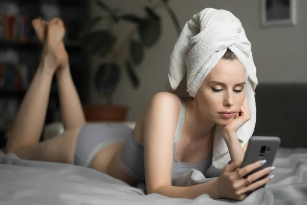 Bella giovane donna con un asciugamano in testa si trova sul telefono cellulare tenere a letto e controllare i social network. Lavoro a distanza, shopping online, isolamento in quarantena — Foto Stock