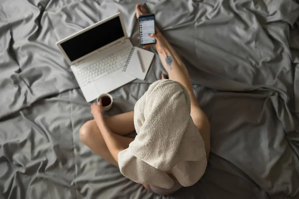 Mujer joven y bonita con una toalla en la cabeza sentada en la cama sostiene el teléfono móvil y revisa las redes sociales. Trabajo remoto en casa con computadora portátil, compras en línea, aislamiento de cuarentena. Vista superior —  Fotos de Stock