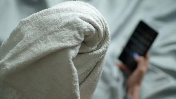 Het meisje gebruikt internet via de telefoon. Vrouw met handdoek op haar hoofd ligt op bed en gebruikt smartphone om nieuwsfeed door te bladeren. Close-up — Stockvideo