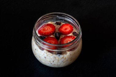 Yarısı çilek ve kuru üzümle dekore edilmiş cam kavanozda bir gecelik yulaflar ortada, açı manzaralı, çatal kaşıklı.