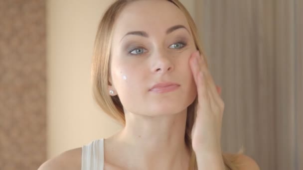 Улыбающаяся молодая женщина наносит крем для лица — стоковое видео
