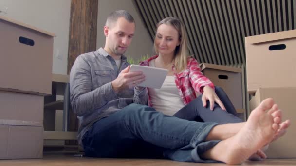 Молодая пара сидит на полу с планшетным ПК и выбирает мебель для своей новой квартиры — стоковое видео