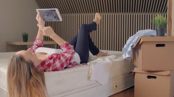 Woman lying on bed using digital tablet instead of unpacking boxes of clothes in new home — Stock Video