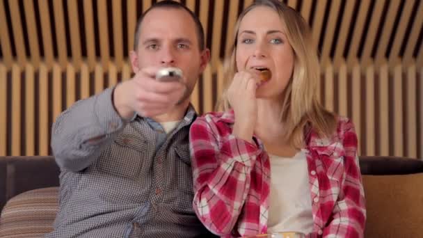 Casal jovem assistindo TV no sofá na sala de estar acabar comendo batatas fritas — Vídeo de Stock