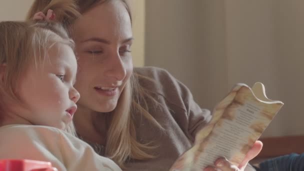 Uśmiechający się matka i córka razem czytając książkę w salonie. — Wideo stockowe