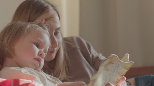 Retrato de una joven y sonriente linda madre e hija leyendo un libro tumbada y relajada en la cama — Vídeos de Stock