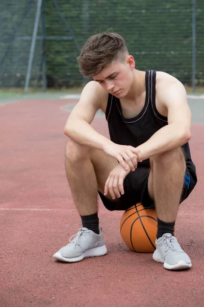 Adolescente sentado em uma bola de basquete em um tribunal parecendo triste — Fotografia de Stock