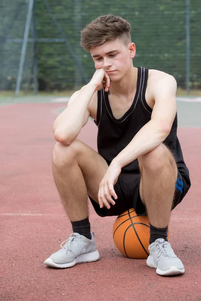 Adolescente sentado em uma bola de basquete em um tribunal parecendo triste — Fotografia de Stock