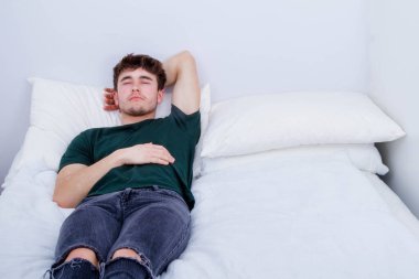 Genç yetişkin bir adam sırt üstü yatar ve elbiseleriyle bir yatakta uyur.