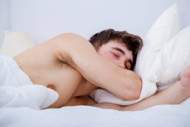 Çıplak göğüslü bir yatakta uyuyan genç bir adam.