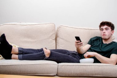 Genç yetişkin bir adam kanepede uzanıp televizyon izliyor ve abur cubur yiyor.