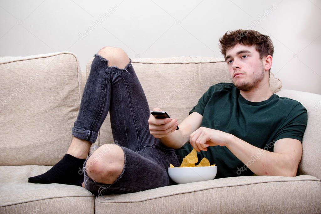 A young adult man laying on a sofa watching TV and eating snacks