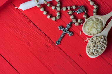 silver Christian cross necklace on white candle,  Bible and seeds of rice and wheat in a wooden spoon - bright red on the old table. as a symbol of the beginning of Great Lent - Toned image  clipart