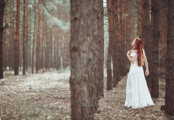 Ginger girl in white dress walking in pine forest.