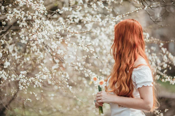 Ginger girl luktar vårblommor nära blommande trädet. Stockbild