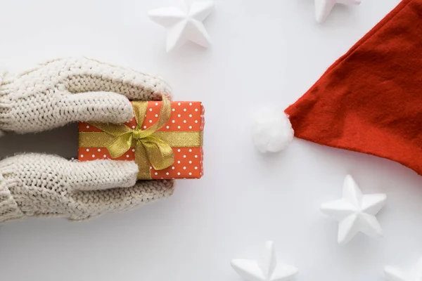 Руки жінки в рукавичках тримають червону подарункову коробку ізольовано на білому тлі з білими зірками та Санта-капелюхом. Вид зверху, плоска лежала. Копіспазмі . — стокове фото