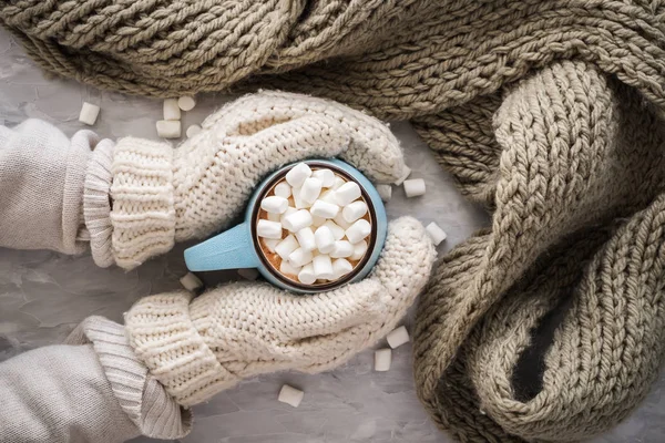 Christmas and New Year cozy holiday composition with scarf, woman hands in mittens, mugs with cocoa or chocolate, marshmallow on the gray concrete background. Flat lay, top view. — Stock Photo, Image