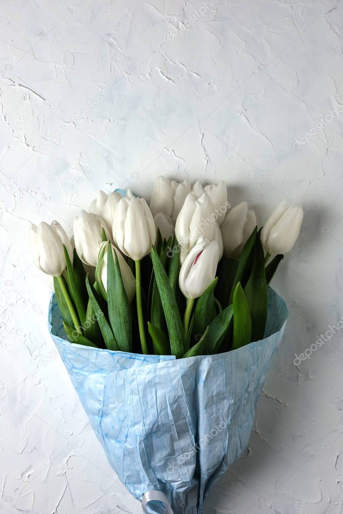 A bouquet of white tulips in blue wrapping paper on a gray concrete background. Top view. Flat lay. Postcard for Easter, Mothers Day and Spring Holidays.