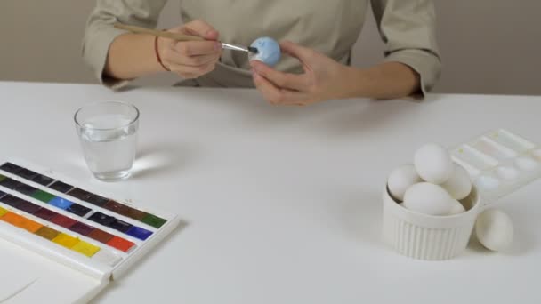 Eine unkenntliche Frau bemalt Ostereier mit einem Pinsel. Auf dem Tisch liegt ein Glas Wasser und eine Schüssel mit Eiern. Vorbereitung auf die Feiertage. — Stockvideo